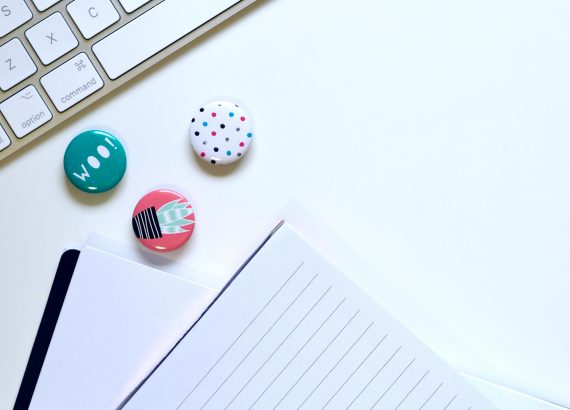 Flat lay of a minimalist workspace featuring a keyboard, colorful badges, and a notepad on a white background.
